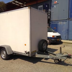 Geschlossener weißer Anhänger mit einem Ersatzreifen an der Deichsel, geparkt auf einem Hof vor blauen Containern